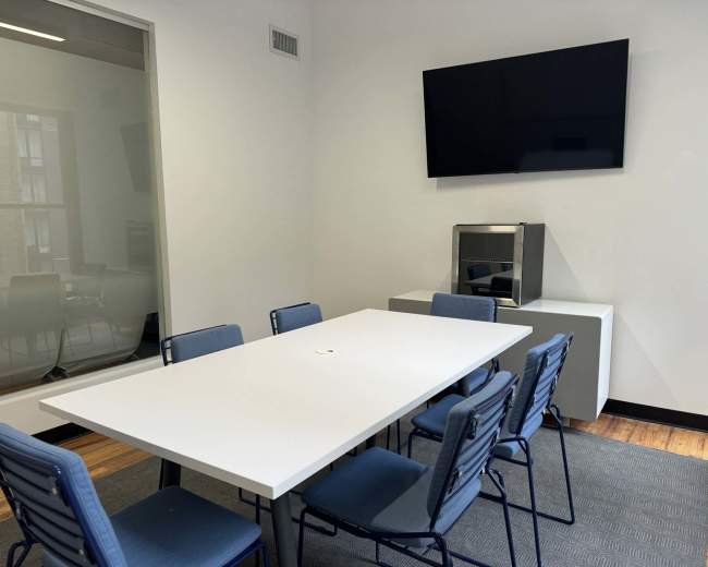 The image shows a conference room with a long white table, blue chairs, a mounted television, and a small cabinet in a minimalist setting.