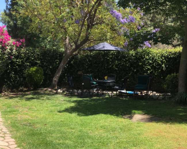 The image shows a grassy backyard with a stone pathway, a shaded seating area under a canopy, and flowering shrubs along the perimeter.