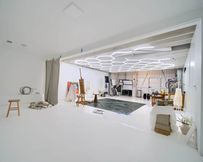 The image shows a spacious white studio with a variety of art supplies and furniture, including a stool, a large rug, and easels, illuminated by a hexagonal ceiling light.