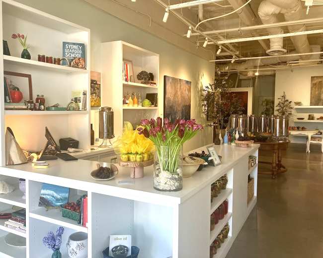 The image shows a bright, modern retail space featuring shelves filled with various food items, decorative objects, and a bouquet of tulips on a counter.