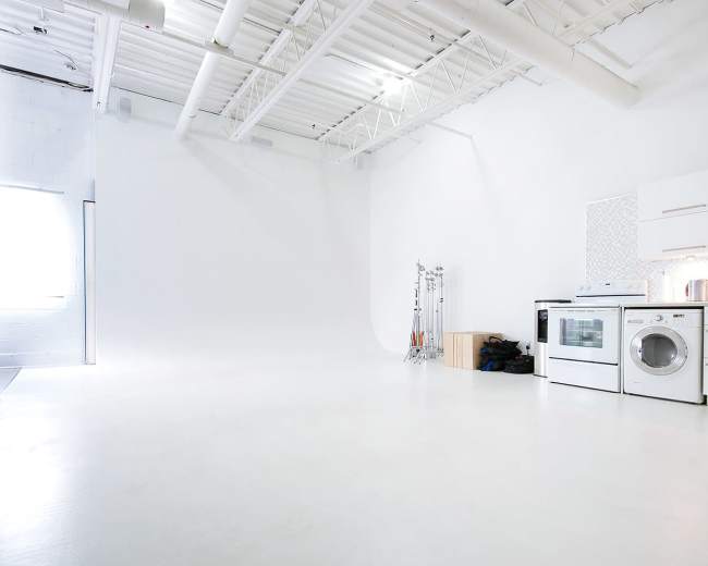 The image shows a spacious, bright studio with white walls and a polished floor, featuring a kitchen area with a stove and washing machine in the corner.