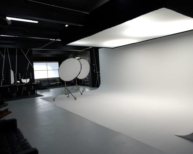 The image shows a photography studio with a large white backdrop, softbox lighting, and reflector tools set up in a minimalist space.