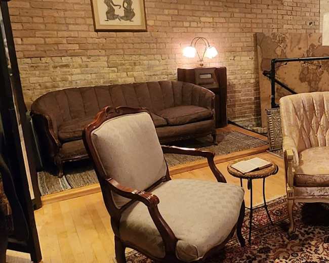 The image shows a cozy living room with a brick wall, featuring a vintage armchair and a patterned area rug surrounded by various seating arrangements and decor items.