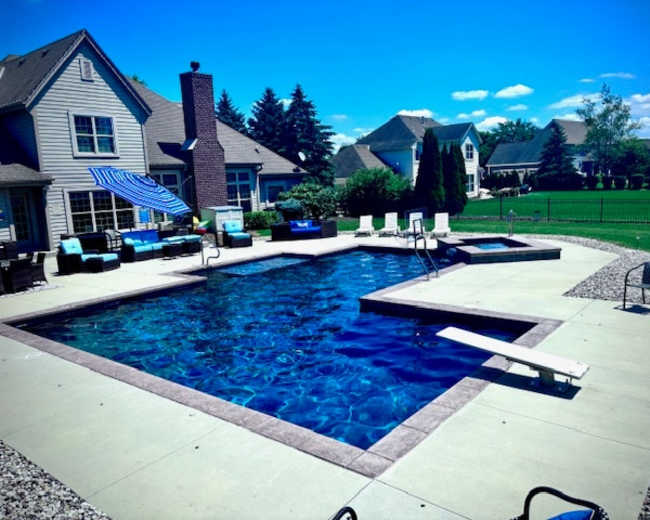 The image shows a residential backyard featuring a swimming pool with a diving board, lounge chairs, and a patio area adjacent to a house.