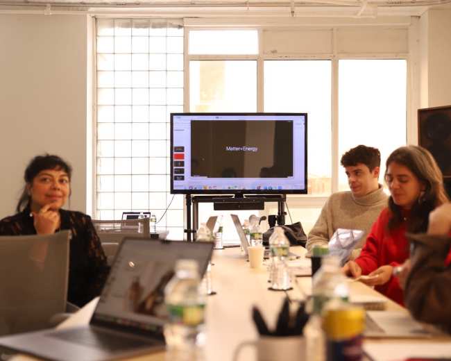 A group of five people is seated around a table during a meeting, with a presentation screen displaying a title in the background.