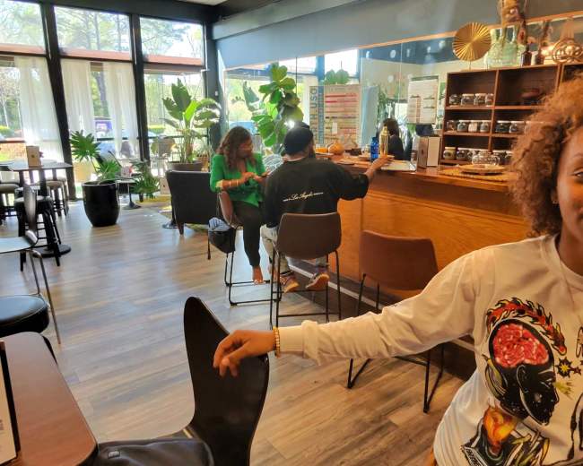 The image shows a cozy café interior with patrons seated at a counter and various plants decorating the space.