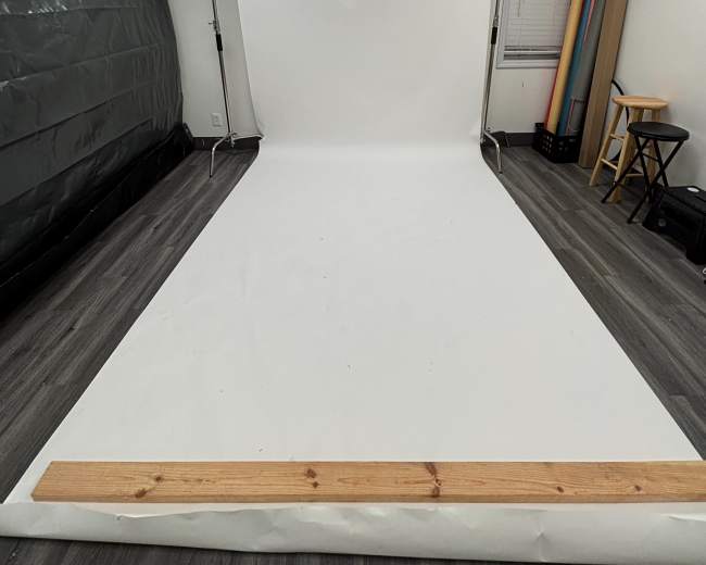 The image shows a photography studio with a white backdrop, wooden flooring, and various backdrop rolls in the corner.
