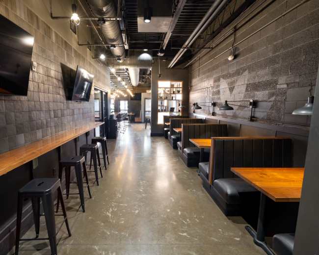 The image shows a modern, narrow restaurant space featuring dark walls, wooden tables, and booth seating along one side.