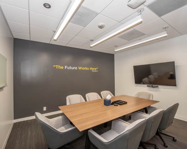 A conference room features a large wooden table surrounded by six gray chairs, with a wall displaying the phrase "The Future Works Here" and a mounted TV.