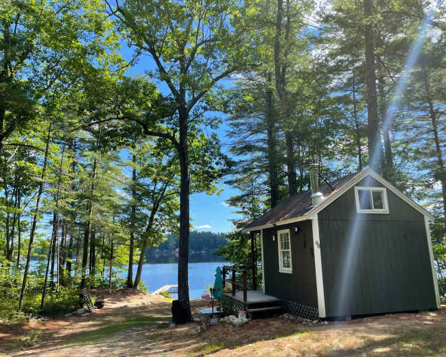 A small green cabin is situated near a lake, surrounded by tall trees and clear blue skies.