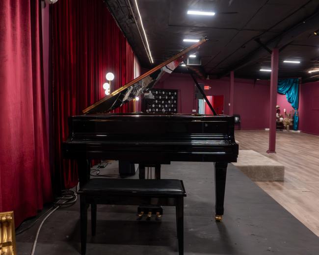 A black grand piano is positioned on a stage in front of red curtains, with a wooden bench nearby and a backdrop of dim lighting.
