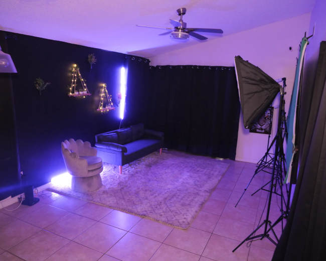 The image shows a well-lit indoor space featuring a black wall with decorative lights, a gray couch, and a large area rug on tiled flooring.