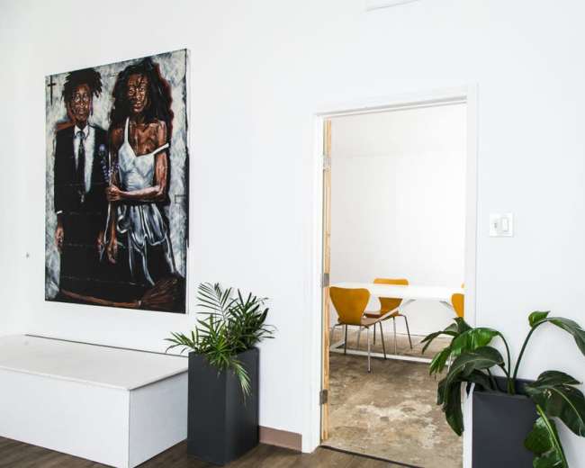 The image shows an interior space with a large painting of two figures on a white wall, a doorway leading to a bright room, and potted plants in the foreground.