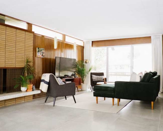 The image shows a modern living room with a dark green sectional sofa, two gray armchairs, a television, and large windows allowing natural light.