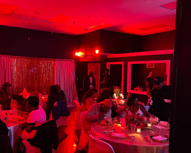 A dimly lit event space with guests seated at elegantly set tables, illuminated by red lighting and decorated with shimmering backdrops.