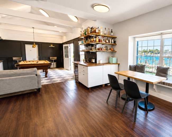 The image shows an interior space with a modern kitchen area, a dining table for two, a seating area with a couch, and large windows overlooking a waterfront with boats.