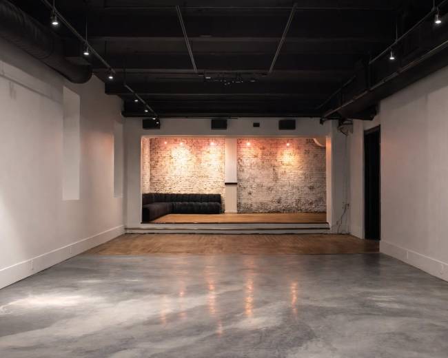 The image shows a spacious, minimalist room with a polished concrete floor, a brick wall with soft lighting, and a sectional couch in a recessed area.