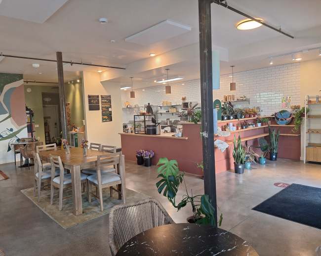 The image shows a modern café interior with a seating area, a service counter, and decorative plants.