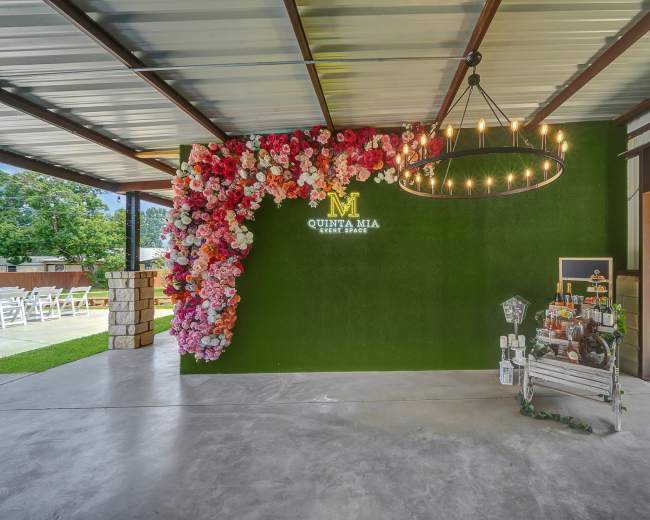 The image shows a decorated event space featuring a floral wall, a chandelier, and a bar cart against a green backdrop.