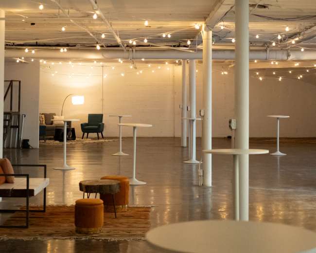 The image shows a spacious interior room with a polished concrete floor, several white tables, and soft lighting from string lights above.
