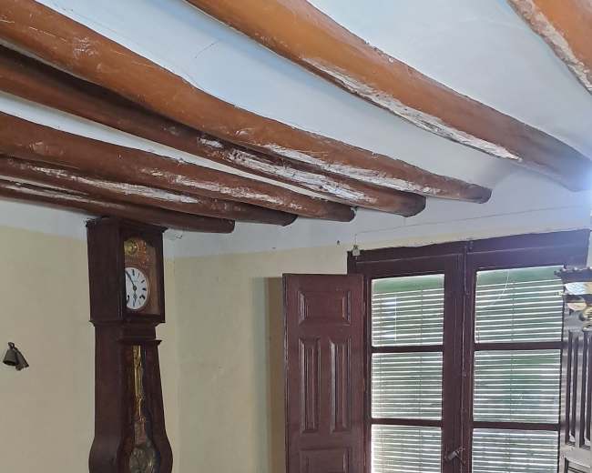 The room features wooden beams on the ceiling, a tall antique clock, two wooden chairs, a table with a pot, and a door leading outdoors.