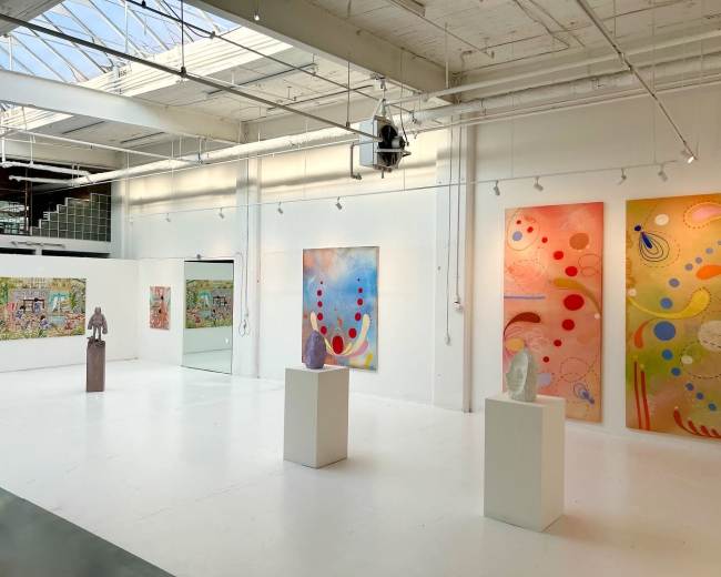 The image shows an art gallery featuring colorful abstract paintings on the walls and sculptures displayed on pedestals in a spacious, well-lit environment.