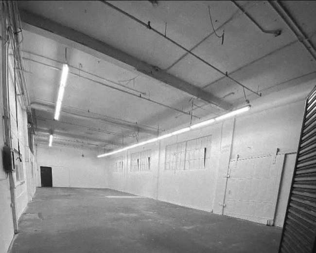 The image shows a large, empty industrial warehouse space with concrete flooring and overhead lighting.