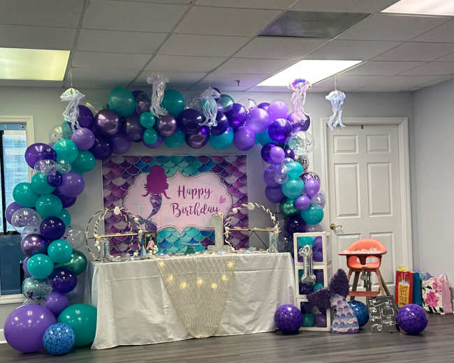 A birthday celebration setup features a decorated table with a "Happy Birthday" banner, surrounded by purple and teal balloons.