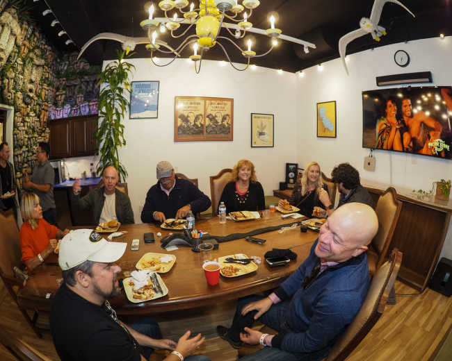 A group of people gathers around a large table, enjoying food and drinks while engaged in conversation in a brightly lit room decorated with various pictures and plants.