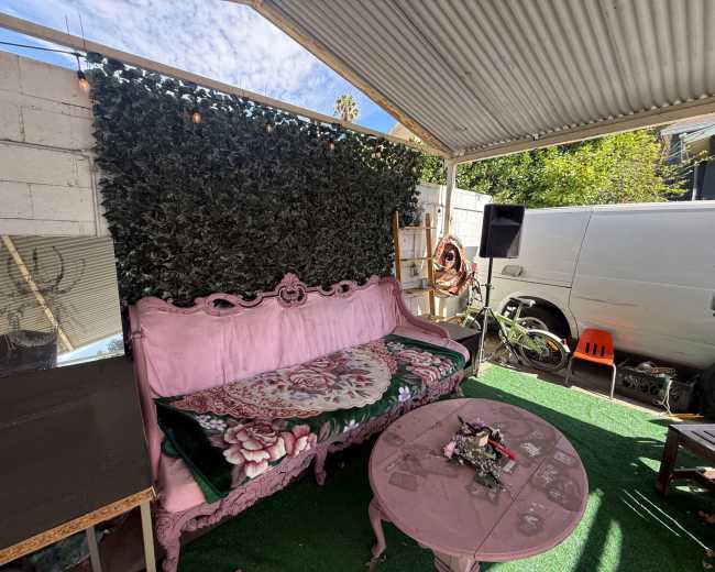 The image shows a cozy outdoor seating area with a pink vintage couch, a round table, and a green artificial grass floor beneath a metal roof.