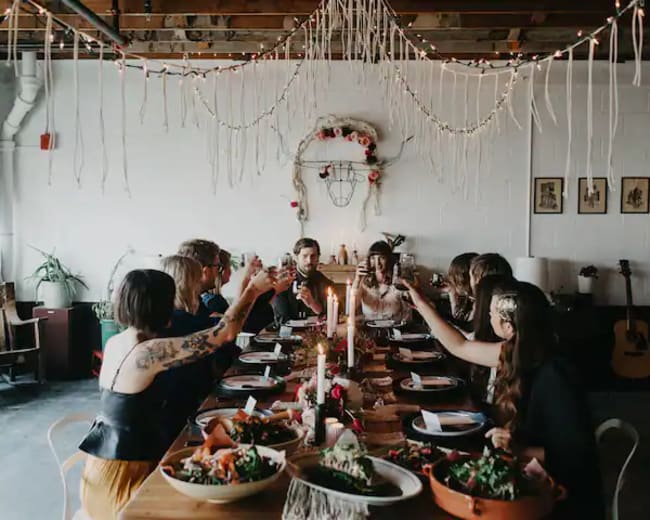 A group of people sits around a long table decorated with candles and food, raising their glasses in a toast.