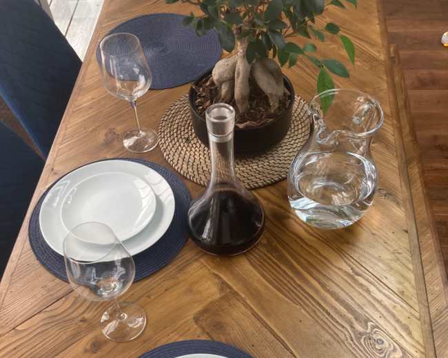A wooden dining table is set with two white plates, wine glasses, a decanter filled with red wine, a pitcher of water, and a potted plant in the center.
