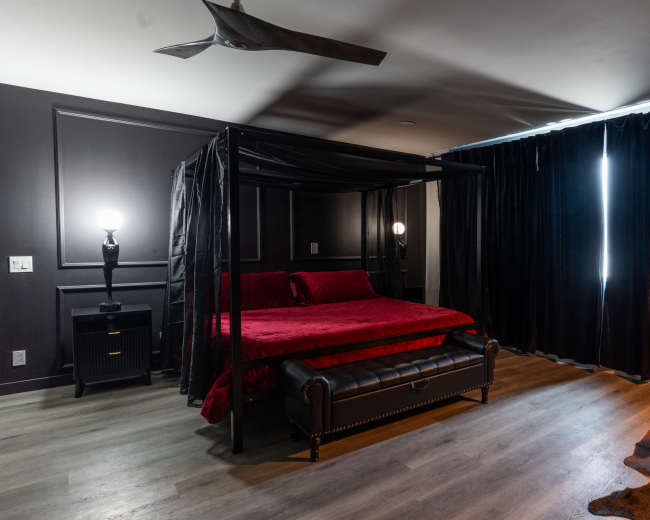 The image shows a dark-themed bedroom featuring a four-poster bed with red bedding, flanked by two lamps, a black leather sofa, and large black curtains.