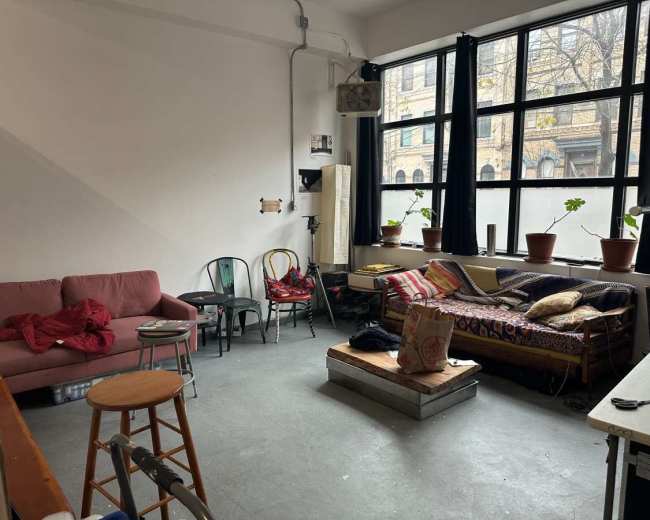 The image shows a partially furnished room with a pink couch, a wooden stool, a patterned sofa, potted plants on windowsills, and a table in the corner.