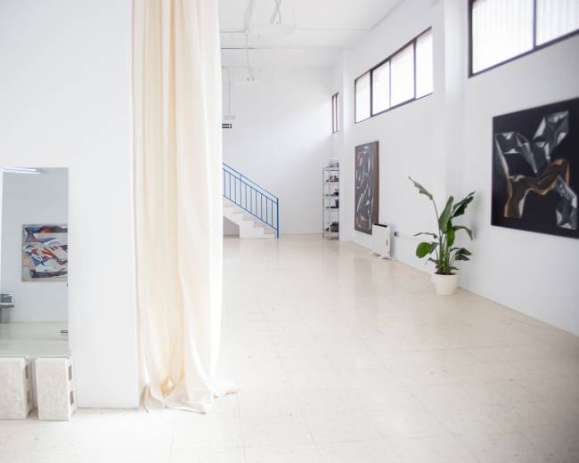The image depicts a spacious, minimalist art studio with white walls, natural light, two artworks hanging on the walls, and a staircase in the background.