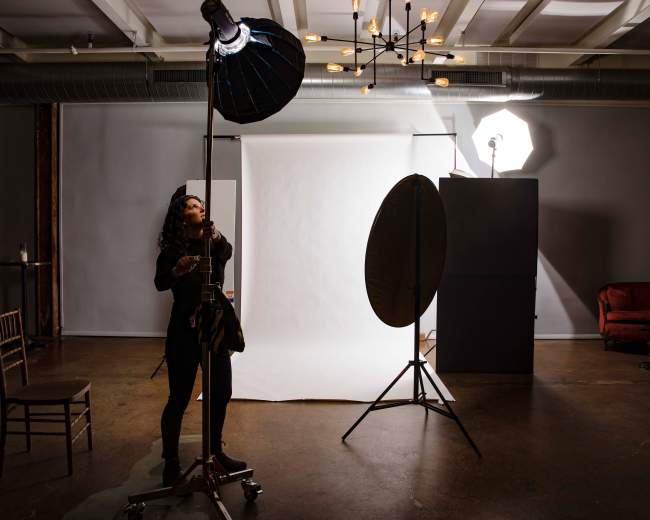 A woman adjusts a photography light in a studio setting with a plain backdrop and various lighting equipment.