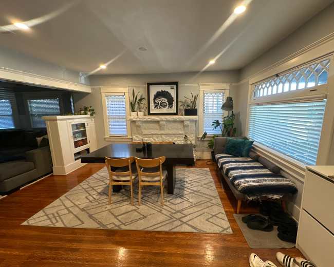 The image shows a cozy dining room with a black dining table, wooden chairs, and a stone fireplace adorned with plants and artwork.