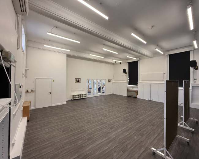 The image shows a large, empty room with wooden flooring, high ceilings, and white walls, featuring windows and mounted light fixtures.