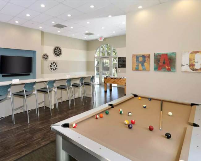 A recreational room with a pool table in the foreground, a bar area with high stools, and a large TV on the wall, decorated with art pieces spelling "RACE."
