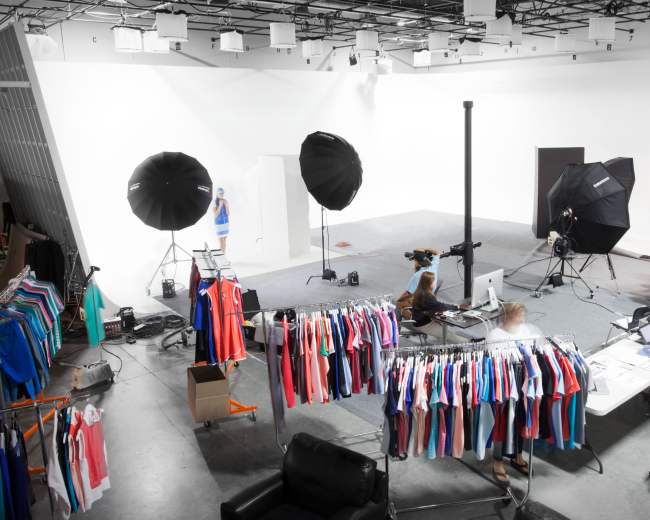 A photography studio features a setup with clothing racks, computer workstations, and multiple lighting sources.