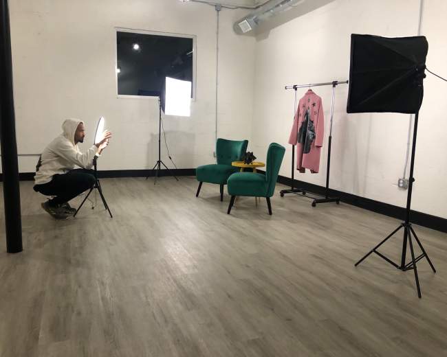 A person in a hoodie is crouched next to a chair in a photography studio, adjusting lighting equipment while a rack with clothing stands nearby.