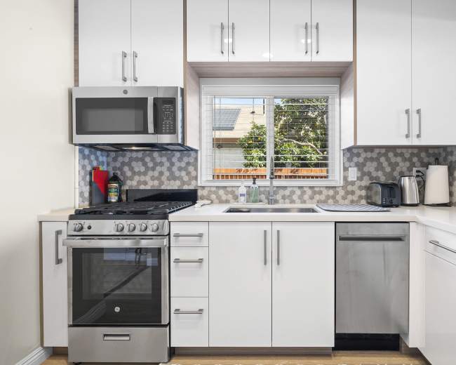 The image shows a modern kitchen featuring stainless steel appliances, white cabinetry, and a window overlooking a garden.