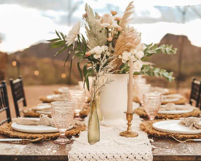 A dining table is set outdoors with elegant dishes, decorative glassware, and a floral centerpiece surrounded by mountains.