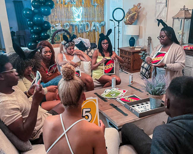A group of people gathers in a living room, sitting around a coffee table with colorful cards while decorations for a birthday celebration are displayed in the background.