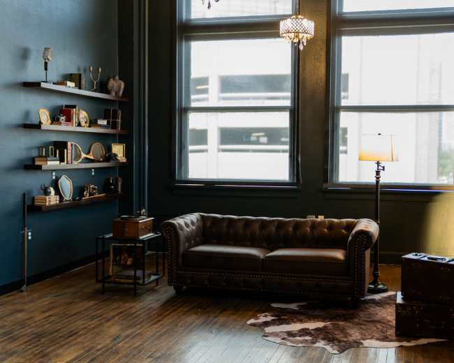 The image shows a cozy room with a brown leather couch, decorative shelves on the walls, a large mirror, a lamp, and two vintage suitcases on the floor.