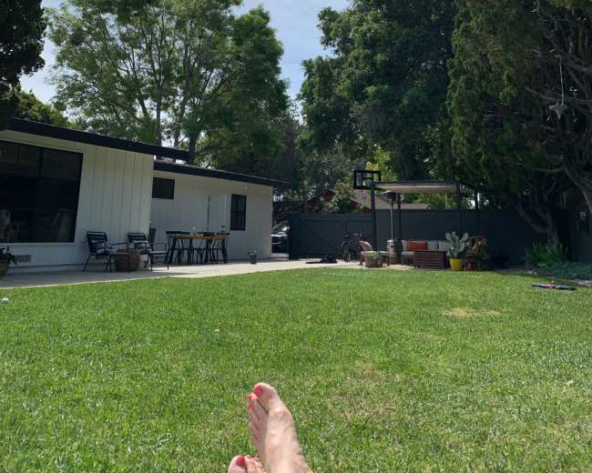 A person is relaxing on a green lawn in a backyard with trees and a house in the background under a partly cloudy sky.