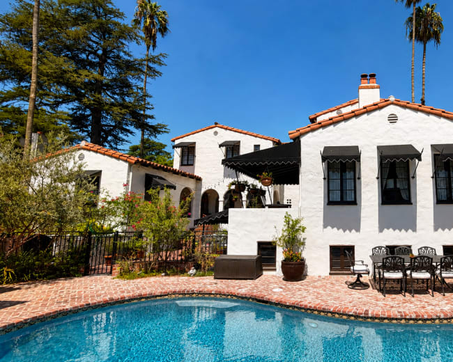 The image shows a white stucco house with a red tile roof beside a swimming pool, surrounded by lush greenery and patio furniture.