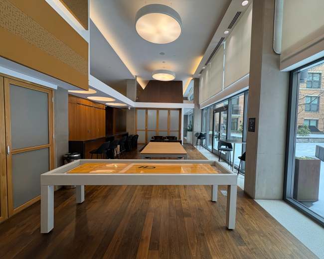 The image shows a modern, spacious interior featuring wooden flooring, contemporary furniture, and large windows overlooking an outdoor area.