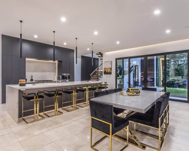 A modern kitchen and dining area features a spacious island with bar stools, sleek cabinetry, and large windows overlooking a garden.
