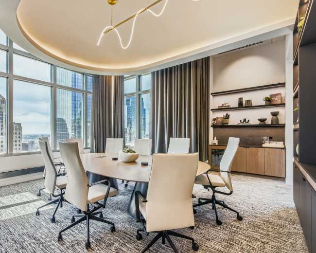 A modern conference room features a circular table surrounded by white ergonomic chairs, with large windows offering a city skyline view.